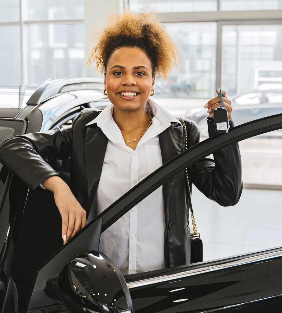 A smiling customer holding her new car key beside an open car door in a dealership showroom